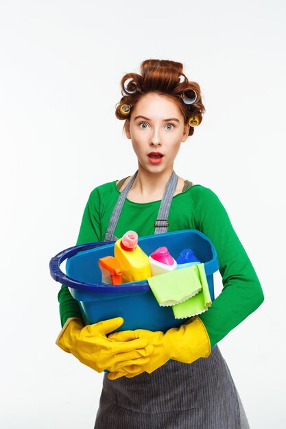 Überraschte nette Frau hält blauen Eimer voller Reinigungswerkzeuge