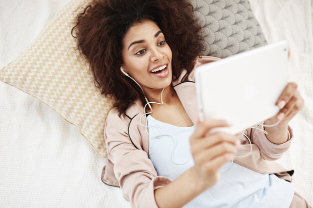 Überraschte schöne afrikanische Frau in den Kopfhörern, die lächelnd betrachten Tablette, die auf Bett liegt. Wachte so auf und machte morgens ein Selfie