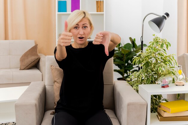 Überraschte schöne blonde russische Frau sitzt auf Sessel Daumen hoch und Daumen runter und schaut auf die Kamera im Wohnzimmer