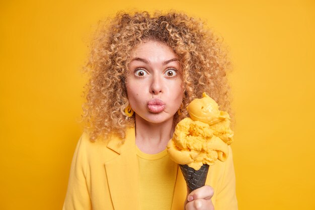 Überraschte schöne Frau hält großes Eis in schwarzer Waffel hält die Lippen gefaltet, fühlt sich in Versuchung, kaltes Sommerdessert in modischer Kleidung isoliert über gelber Wand zu essen