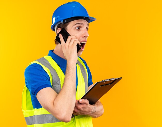 Überraschter Blick auf die Seite junger Baumeister in Uniform mit Zwischenablage spricht am Telefon isoliert auf gelber Wand mit Kopierraum