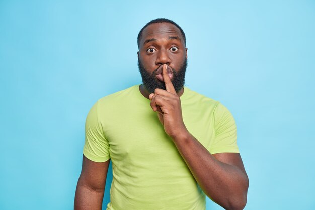 Überraschter erwachsener Mann sagt, Shh bittet, ruhig zu sein, schockiert zu sein, wenn Gerüchte den Finger auf die Lippen drücken, trägt grünes T-Shirt isoliert über blauer Wand
