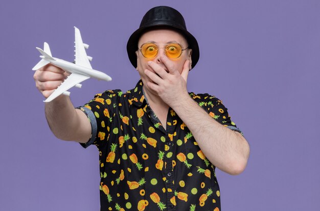 Überraschter erwachsener slawischer Mann mit schwarzem Hut mit Sonnenbrille, der die Hand auf den Mund legt und das Flugzeugmodell hält