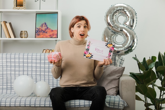 Überraschter gutaussehender Kerl am glücklichen Frauentag, der Geschenk mit Grußkarte auf dem Sofa im Wohnzimmer hält