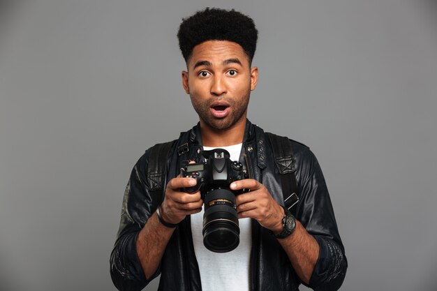 Überraschter hübscher afroamerikanischer Mann in der Lederjacke, die Digitalkamera hält