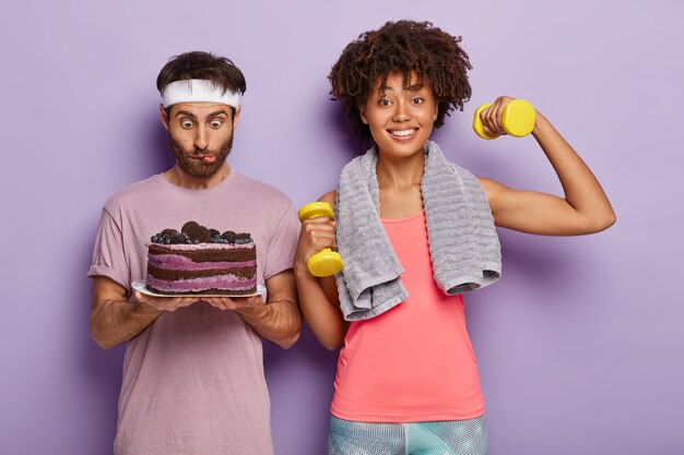 Überraschter junger Afro-Mann starrt auf köstlichen Kuchen, trägt weißes Stirnband, fühlt Versuchung, glückliche Frau arbeitet am Bizeps, erhöht Gewichte, führt sportlichen Lebensstil, steht vor lila Hintergrund.