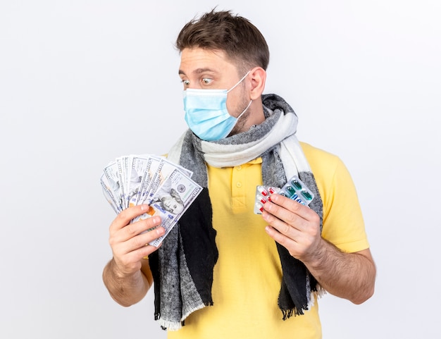 Überraschter junger blonder kranker Mann, der medizinische Maske und Schal trägt, hält Geld und Packungen von medizinischen Pillen, die auf weißer Wand lokalisiert werden