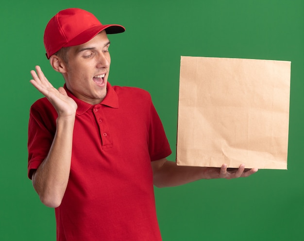 Überraschter junger blonder Lieferjunge steht mit erhobener Hand, die Papierpaket einzeln auf grüner Wand mit Kopienraum hält und betrachtet