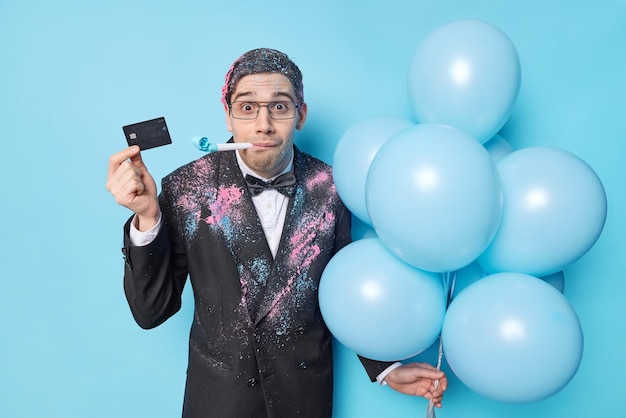 Überraschter junger erwachsener Mann in festlicher Kleidung bläst Partyhorn hält Kreditkarte und Haufen aufgeblasener Luftballons feiert besonderen Anlass isoliert über blauem Hintergrund Urlaubskonzept