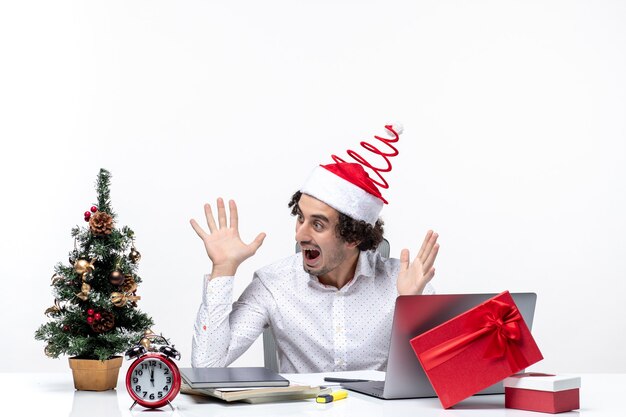 Überraschter junger Geschäftsmann mit lustigem Weihnachtsmannhut, der Weihnachtsbaum verziert und Weihnachten im Büro auf weißem Hintergrund feiert