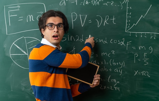 Überraschter junger kaukasischer Geometrielehrer, der eine Brille trägt, die in der Profilansicht vor der Tafel im Klassenzimmer steht und eine Mini-Tafel hält, die Kreide auf der Tafel mit Blick auf die Seite zeigt