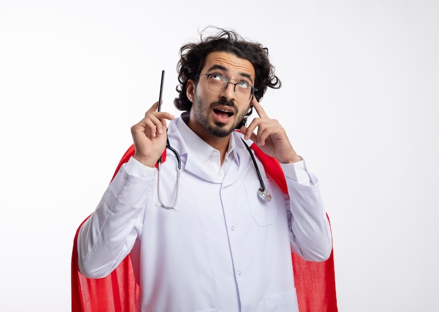 Überraschter junger kaukasischer Superheldenmann in der optischen Brille, die Arztuniform mit rotem Umhang und mit Stethoskop um Hals trägt, schaut und zeigt mit Bleistift mit Kopienraum
