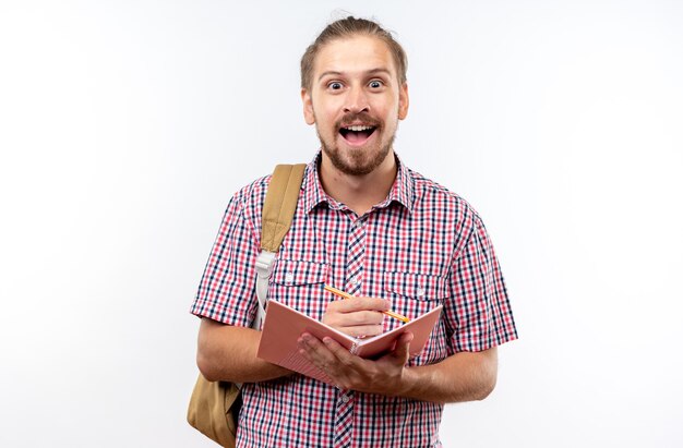 Überraschter junger Kerl, der einen Rucksack trägt und etwas auf ein Notizbuch schreibt, das auf weißer Wand isoliert ist?