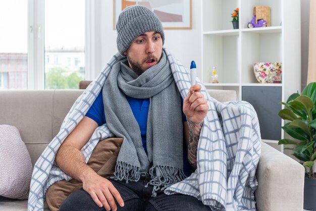 Überraschter junger kranker Mann mit Schal und Wintermütze, der in Decke gewickelt auf dem Sofa im Wohnzimmer sitzt und die Hand am Bein hält und auf das Thermometer schaut
