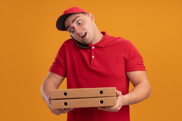 Überraschter junger Lieferbote, der Uniform mit Kappe hält, die Pizzaschachteln hält, spricht am Telefon lokalisiert auf orange Wand