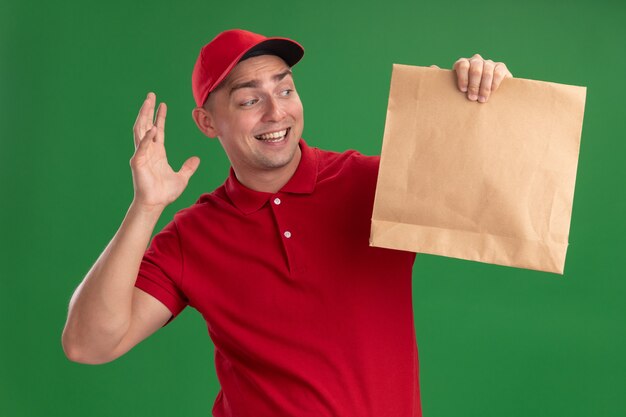 Überraschter junger Lieferbote, der Uniform und Kappe hält und Papiernahrungsmittelpaket lokalisiert auf grüner Wand hält