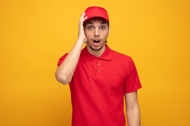 Überraschter junger Lieferbote in Uniform und Mütze, der die Hand auf dem Kopf hält und isoliert auf gelbem Hintergrund in die Kamera blickt