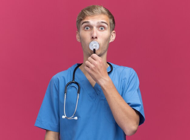 Überraschter junger männlicher Arzt, der Arztuniform mit Stethoskop trägt, das Stethoskop auf Mund lokalisiert auf rosa Wand setzt