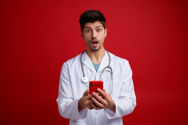 Überraschter junger männlicher Arzt, der eine medizinische Uniform und ein Stethoskop um den Hals trägt und ein Mobiltelefon mit beiden Händen hält, die isoliert auf rotem Hintergrund in die Kamera blicken