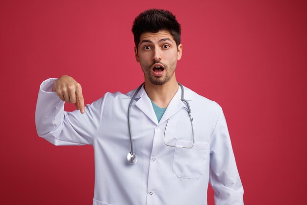 Überraschter junger männlicher Arzt, der medizinische Uniform und Stethoskop um den Hals trägt und in die Kamera blickt, die isoliert auf rotem Hintergrund nach unten zeigt