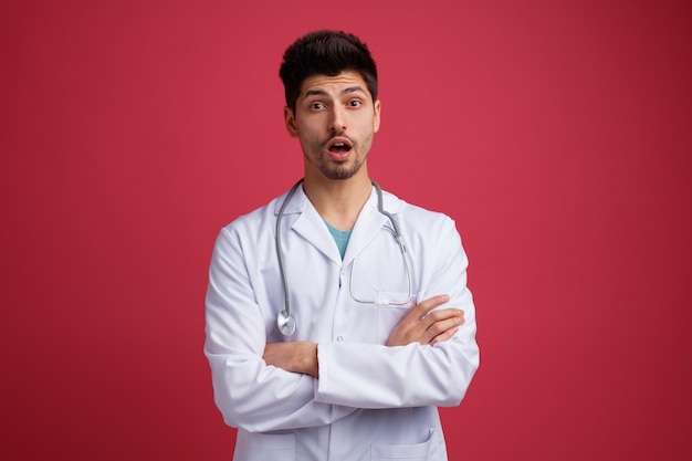 Überraschter junger männlicher Arzt mit medizinischer Uniform und Stethoskop um den Hals, der in die Kamera schaut, während er die Arme isoliert auf rotem Hintergrund verschränkt hält