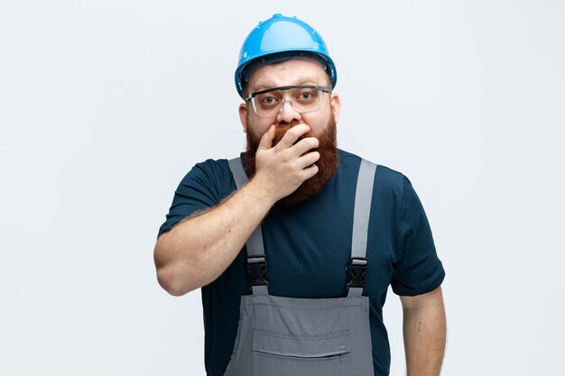 Überraschter junger männlicher Bauarbeiter mit Schutzhelmuniform und Schutzbrille, der die Hand auf dem Mund hält und isoliert auf weißem Hintergrund in die Kamera blickt