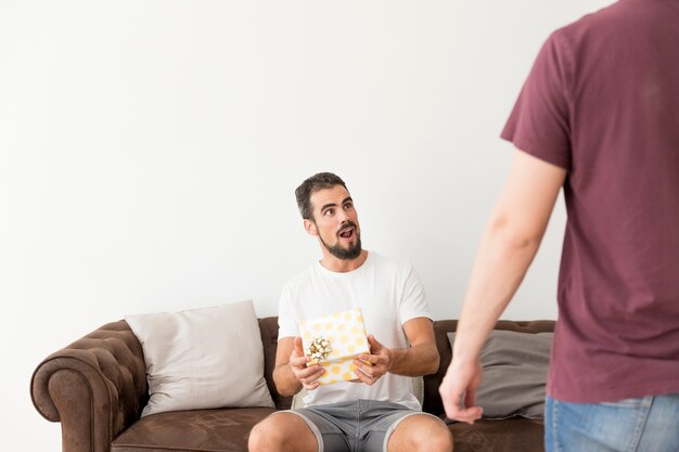 Überraschter junger Mann, der auf dem Sofa hält die Geschenkbox betrachtet seinen Freund sitzt