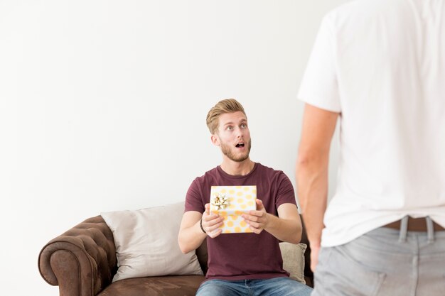 Überraschter junger Mann, der auf dem Sofa hält die Geschenkbox betrachtet seinen Freund sitzt