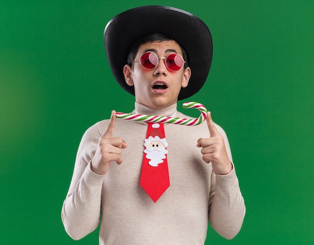 Überraschter junger Mann, der Hut mit Weihnachtskrawatte und Brille trägt, die Weihnachtsbonbons lokalisiert auf grünem Hintergrund hält