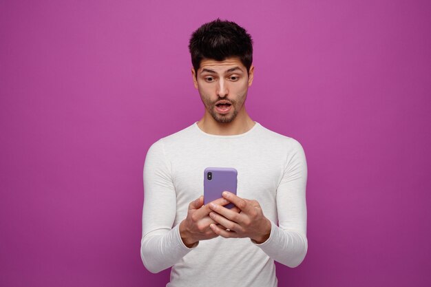 Überraschter junger Mann mit seinem Handy isoliert auf violettem Hintergrund