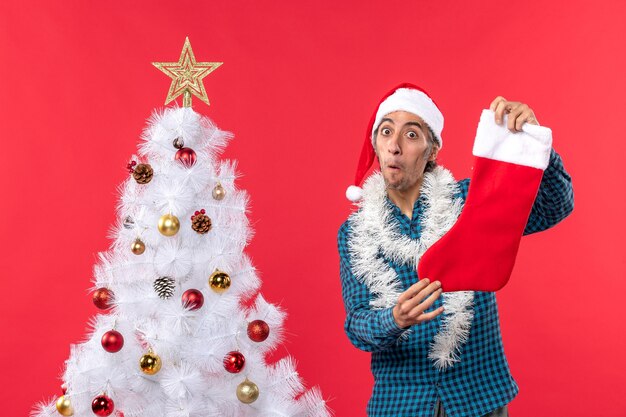 Überraschter junger Mann mit Weihnachtsmannhut in einem blauen gestreiften Hemd und Halten Weihnachtssocke nahe Weihnachtsbaum auf Rot