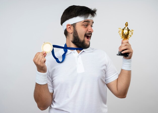 Überraschter junger sportlicher Mann, der Stirnband und Armband mit Halten und Betrachten des Siegerpokals trägt und Medaille trägt