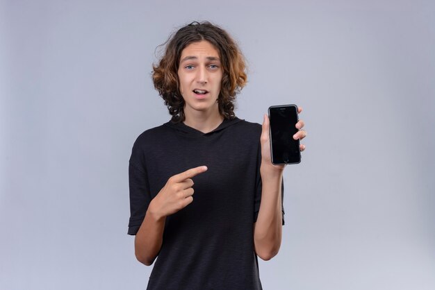 Überraschter Kerl mit langen Haaren im schwarzen T-Shirt, der ein Telefon hält und auf Telefon auf weißem Hintergrund zeigt