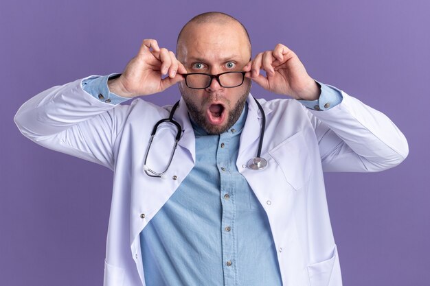 Überraschter männlicher Arzt mittleren Alters, der ein medizinisches Gewand und ein Stethoskop mit einer Brille trägt, die eine Brille greift und nach vorne isoliert auf lila Wand schaut