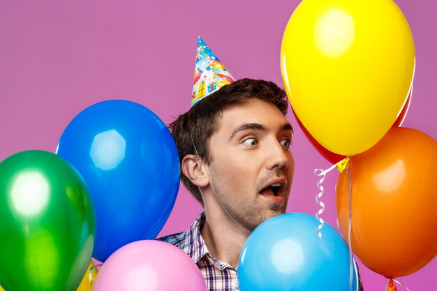 Überraschter Mann, der Geburtstag feiert und bunte Luftballons über lila Wand hält.