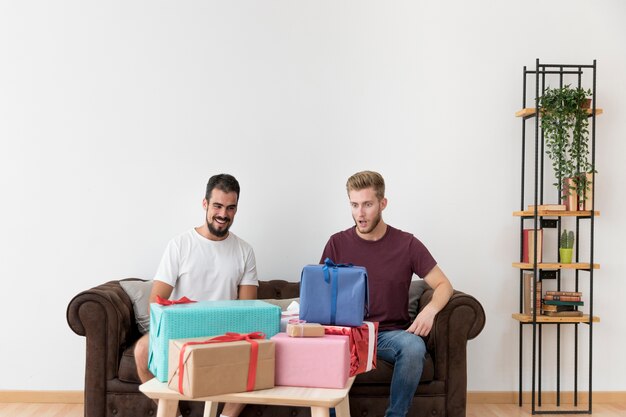 Überraschter Mann, der viele bunten Geschenkboxen sitzen mit seinem Freund betrachtet