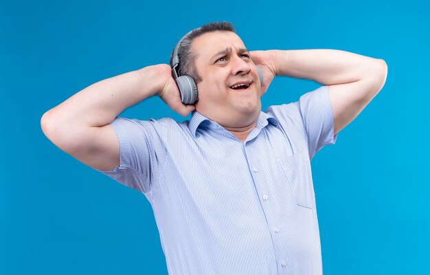 Überraschter Mann des mittleren Alters im blau gestreiften Hemd, das Kopfhörer trägt, die Musik auf einem blauen Hintergrund genießen