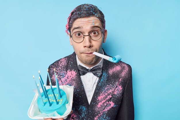 Überraschter Mann feiert Jahrestag bläst Geburtstagshorn hält Kuchen mit brennenden Kerzen in festlicher Kleidung gekleidet hat sich über blauen Hintergrund isolierter Ausdruck gewundert Konzept für besondere Anlässe