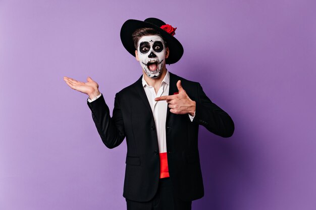Überraschter Mann in der formellen Kleidung, die mit Zombie-Make-up aufwirft. Kaukasischer Typ, der sich auf Halloween im mexikanischen Stil vorbereitet.