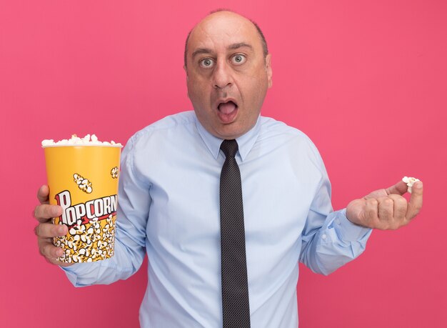 Überraschter Mann mittleren Alters mit weißem T-Shirt mit Krawatte mit Eimer Popcorn und Popcornstück isoliert auf rosa Wandcorn