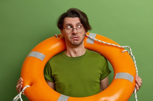 Überraschter Mann posiert mit Bojenring um den Hals, schaut misstrauisch zur Seite, lernt schwimmen, trägt transparente Brille und grünes T-Shirt, wird aus dem Wasser gerettet,