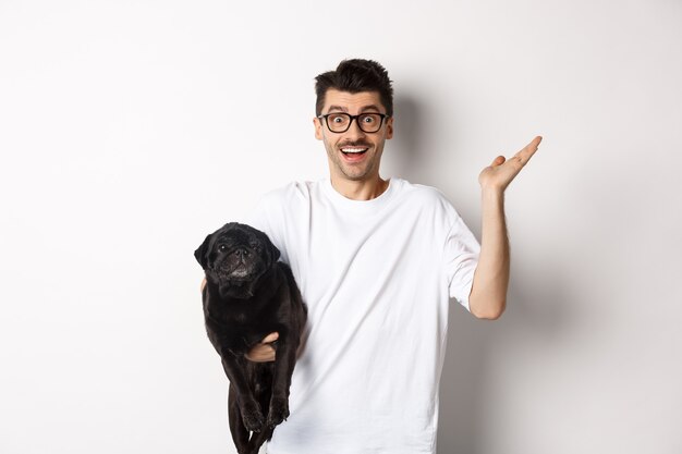 Überraschter und glücklicher Hundebesitzer, der einen süßen schwarzen Mops hält, die Hand erstaunt hebt, zufrieden in die Kamera starrt, auf weißem Hintergrund steht