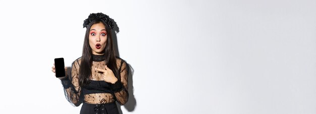 Überraschtes asiatisches Mädchen im schwarzen Gothic-Kleid mit Kranz, das amüsiert nach Luft schnappt und mit dem Finger auf das Handy zeigt