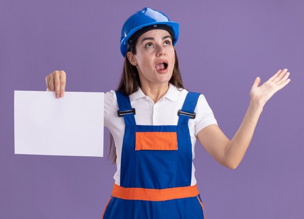 Überraschtes Betrachten der jungen Baumeisterfrau in Uniform, die Papierverbreitungshand lokalisiert auf purpurroter Wand hält
