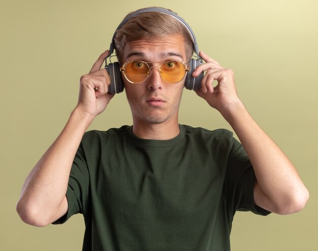 Überraschtes Betrachten des jungen gutaussehenden Mannes des vorderen tragenden grünen Hemdes mit Brille und Kopfhörern lokalisiert auf olivgrüner Wand