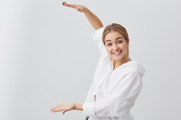 Überraschtes blondes Mädchen in der Freizeitkleidung, die mit Handlänge der Schachtel zeigt. Schöne Frau, die weißes Hemd trägt, mit Zähnen lächelt und Größe von etwas Großem demonstriert. Aktiv gestikulieren