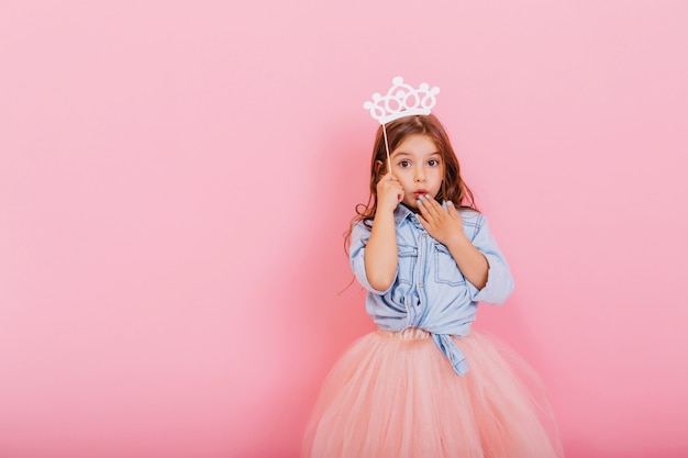 Überraschtes hübsches junges Mädchen im Tüllrock mit Krone auf Kopf, die lokal auf rosa Hintergrund ausdrückt. Erstaunliche süße kleine Prinzessin am Karneval. Platz für Text