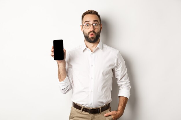 Überraschtes hübsches Manager in der Brille, das neugierig aussieht und mobilen Bildschirm zeigt, der über weißem Hintergrund steht.