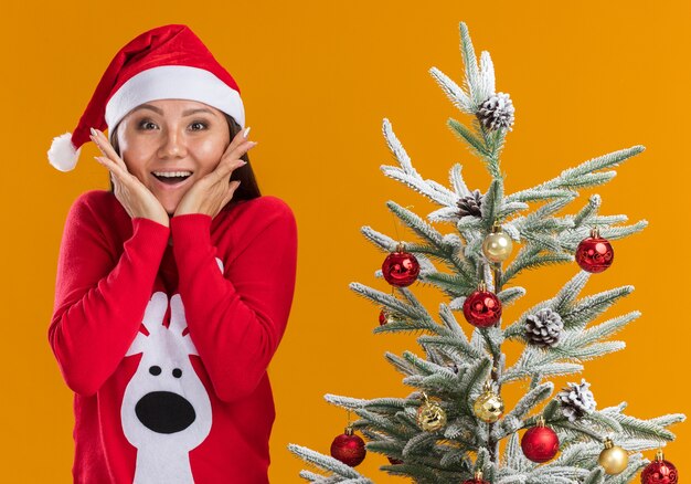 Überraschtes junges asiatisches Mädchen, das Weihnachtshut mit Pullover trägt, der nahe Weihnachtsbaum bedeckte Mund mit Händen lokalisiert auf orange Hintergrund steht