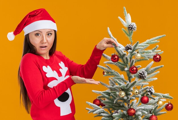 Überraschtes junges asiatisches Mädchen, das Weihnachtsmütze mit Pullover trägt, verziert Weihnachtsbaum lokalisiert auf orange Wand